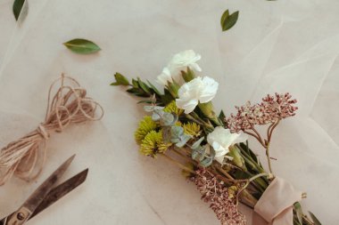 Beyaz karanfilli küçük el yapımı çiçek aranjmanı yeşil kasımpatı, arka planda pembe dantelli okaliptüs yaprakları.