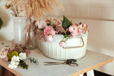 Çiçekçinin çalışma masası. Pembe şakayıklar, beyaz karanfiller, okaliptüs yaprakları, kasımpatı, makas ve vazolarla dolu beyaz masa. Evde buketler düzenleniyor.