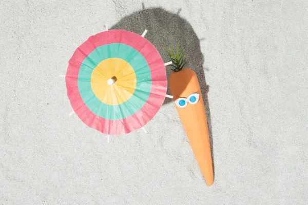 Creative Easter composition with carrots, glasses and beach umbrella on the sand. Summer and holiday idea. Flat lay.