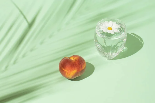 Creative summer composition with fresh peach fruits, a glass of drink and flowers against a pastel green background. Minimal food concept.