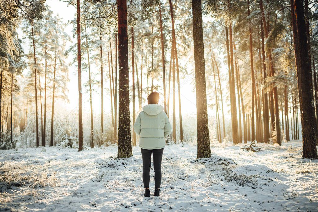 A female in winter hat and puffer standing and observing the beautiful sunset over snow-covered forest