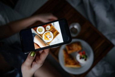 Yatağındaki tahtada bir fincan kahve ve lezzetli hamur işlerinin fotoğrafını çeken bir kadın.