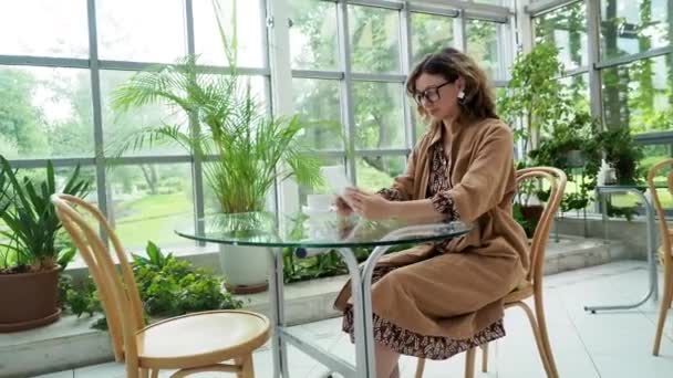 Donkerharige jonge zakenvrouw draagt bril maakt gebruik van moderne tablet zitten aan tafel in lichte tropische pot planten cafe — Stockvideo