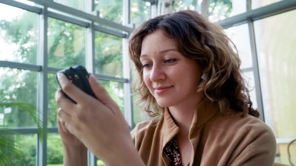 Felice scuro capelli ricci donna d'affari naviga internet con il telefono cellulare seduto a tavola in un ampio caffè. Da vicino. — Video Stock