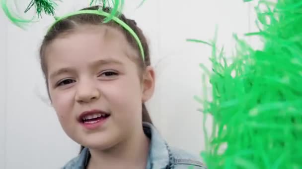 De cerca. Adorable niño feliz pelo oscuro con cuernos divertidos en la cabeza mirando a la cámara, sonriendo y celebrando santo patricks día ondeando con pompones verdes fondo de la pared blanca — Vídeo de stock