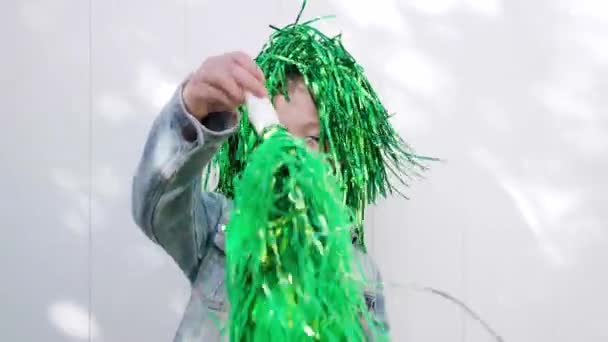 Χαριτωμένο χαρούμενο καστανά μάτια παιδί με αστεία πράσινη περούκα στο κεφάλι της χαμογελώντας στην κάμερα, πηδώντας και γιορτάζοντας την ημέρα του Αγίου Πατρικίου κυματίζει πράσινο πομ πομ λευκό φόντο στον τοίχο — Αρχείο Βίντεο