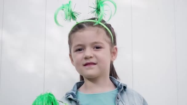 Bela criança feliz cabelos escuros e olhos castanhos com chifres engraçados na cabeça olhando para a câmera, sorrindo e celebrando dia patricks santo acenando com pom verde poms fundo parede branca — Vídeo de Stock