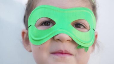 Yeşil karnaval maskeli, yıldızlı şirin kahverengi gözlü kız kameraya bakıyor, gülümsüyor, Aziz Patrick gününü kutluyor, beyaz duvar arkası. Çok yakın çekim.