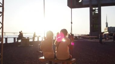 Ağır çekim. Altın bir akşamda sallanırken gün batımını izleyen iki çocuk.