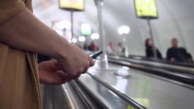 Yürüyen merdivende cep telefonuyla mesaj atan ve kaldıran bir kadın. Diğer insanların metroya inerken arka planları var. Kapat.