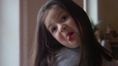 Cute preschooler little kid girl with long brown hair makes a face with sticking out tongue and smiling then. Close up