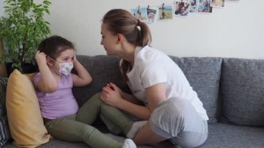 Girl wearing medical mask, her mother helps sitting on homes sofa. Quarantine