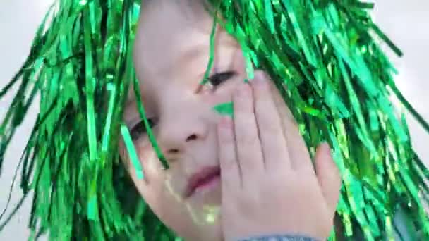 Adorável criança olhos castanhos felizes em peruca verde olhando para a câmera, escondendo estrela verde em sua bochecha, celebrando dia patricks santo, fundo da parede branca. Fechar — Vídeo de Stock