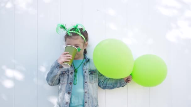 Μικρό παιδί με πράσινα αστεία κέρατα και αστέρι του καρναβαλιού, κρατά δύο πράσινα μπαλόνια, πίνει τσάι από χάρτινο κύπελλο και κοιτάζει την κάμερα, γιορτάζοντας την ημέρα του Αγίου Πατρικίου. Λευκό φόντο τοίχου — Αρχείο Βίντεο
