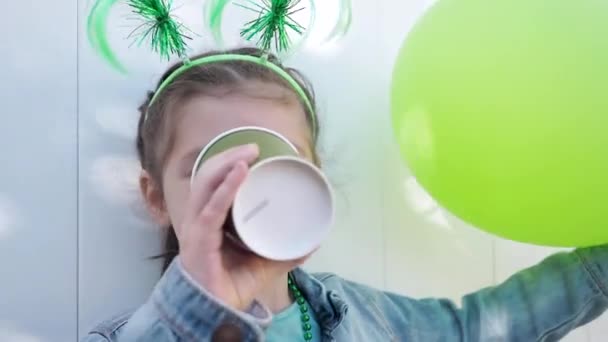 Närbild. Liten unge med gröna roliga horn och karneval ansiktsmask, håller gröna ballonger, dricker te från grön pappersmugg och tittar på kameran, firar St Patricks dag. vit vägg bakgrund — Stockvideo