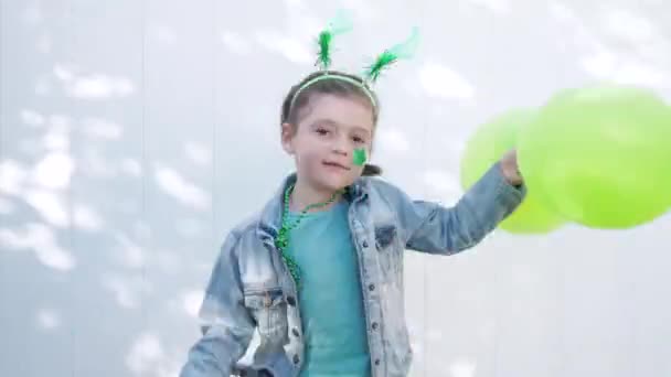 Dziewczyna z zielonymi, zabawnymi rogami na głowie i gwiazdą na policzku trzyma dwa zielone balony i pompony, skacze i uśmiecha się patrząc w obiektyw, świętując dzień świętego Patryka. Białe tło ściany — Wideo stockowe