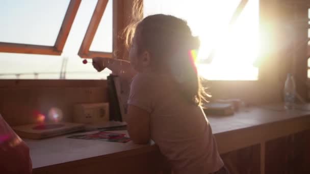 Onherkenbaar klein meisje eten snoep in de voorkant van open raam bij zonsondergang — Stockvideo