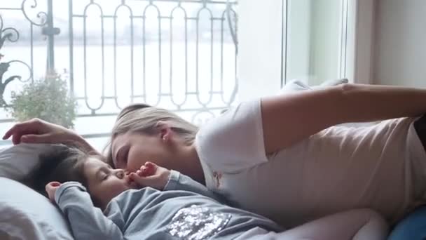 Adorable mother smiling and kissing her sleeping cute preschooler little girl to cheek during nap time in front of window. A kid is awake and smiling — Stok video