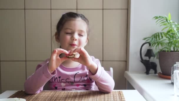 Bedårande fyra år liten flicka njuter av att äta sin mellanmål smörgås med god aptit sitter vid det vita bordet. — Stockvideo