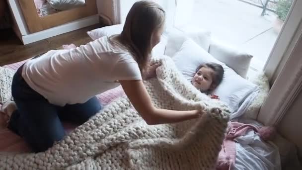 Schattige moeder bedekt met een deken haar schattige kleuter kleine meisje, kust op de wang en vertelt een fee staart, liggend op het bed. Een kind lacht en kijkt naar moeder — Stockvideo