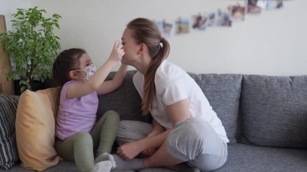 Coronavirusmers. Leuke dochter helpt haar moeder om een medisch masker te dragen terwijl ze thuis op de bank zit. Concept van coronavirus COVID-19 pandemische ziektesymptomen. Home quarantaine. — Stockvideo