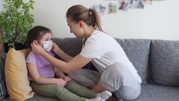 Coronavirus mers. Mère aide sa fille mignonne à porter un masque médical assis sur le canapé à la maison. Concept de coronavirus COVID-19 symptômes de la maladie pandémique. Quarantaine à domicile. — Video