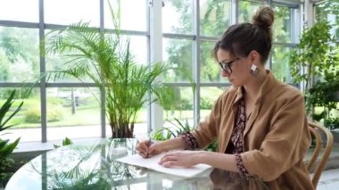 smart serious joung brown hair student with eyeglasses in brown cloak writes in paper notebook with black pen working, earphones at glass table in cafe with tropical flowers during the day