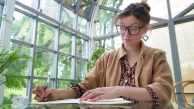 bottom view. smart serious joung brown hair student in brown cloak writes in paper notebook with black pen working, earphones at glass table in cafe with tropical flowers during the day