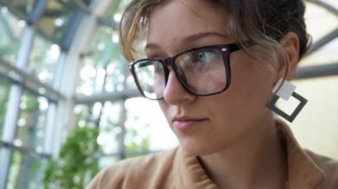 Close up. attractive freelance dark hair businesslady with eyeglasses in brown cloak writes in paper notebook with black pen working in cafe with tropical flowers during the day