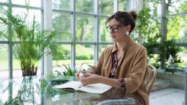 serious beatiful businesslady with dark hair and modern eyeglasses in brown cloak and dress puts on earphones to listen music on smartphone at table in cozy light green cafe