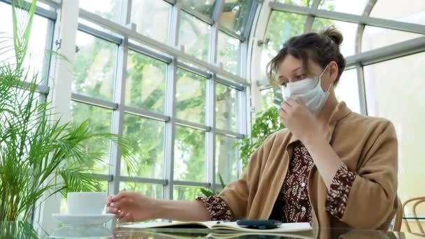 Encantadora dama de pelo oscuro rizado se quita la máscara protectora para beber café fresco en la cafetería con plantas tropicales. — Vídeos de Stock