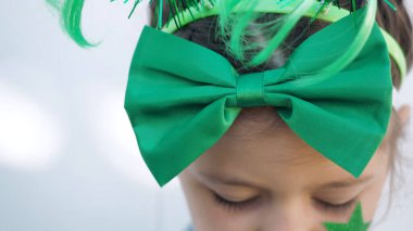 View from above. Cute brown hair girl with big green bow and star on cheek looking down, white wall background, celebrating saint patricks day. Close up