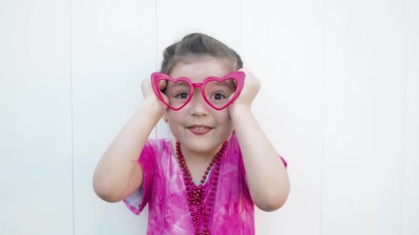 Naughty dark hair girl in funny red Valentines heart glasses, beads and pink dress makes funny silly faces at the camera — Stock Footage Naughty dark hair girl in funny red Valentines heart glasses, beads and pink dress makes funny silly faces at the camera Stock Footage