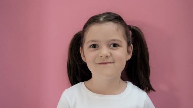 Siyah saçlı ve kahverengi gözlü iki komik at kuyruklu kız gülümseyip kameraya bakıyor. Pembe arkaplan