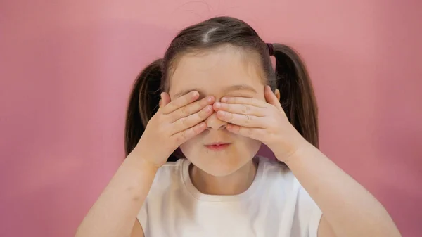 Siyah saçlı şirin bir kızın portresi. İki komik at kuyruklu, beyaz tişörtlü, gözlerini elleriyle kapatan. Pembe arkaplan