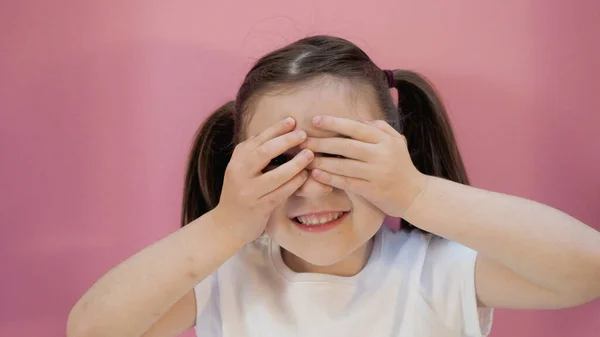 Beyaz tişörtlü güzel bir çocuk iki at kuyruklu gözlerini eliyle kapatıyor ve mutlu bir yüz ifadesi takınıyor. Pembe arkaplan