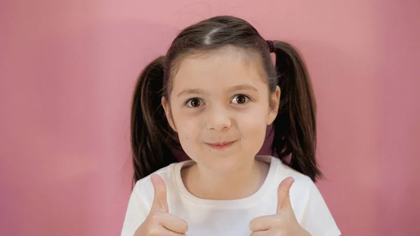 Cute dark hair girl with two funny ponytails shows thumbs up class, smiles, looks at the camera. Pink background