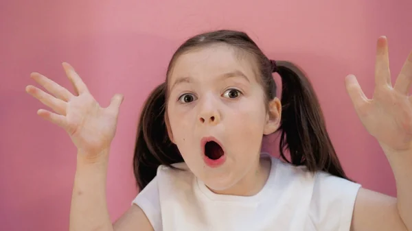 Beyaz tişörtlü, siyah saçlı, at kuyruklu kahverengi gözlü küçük kız şaşırmış elleri kaldırıyor. Pembe arkaplan
