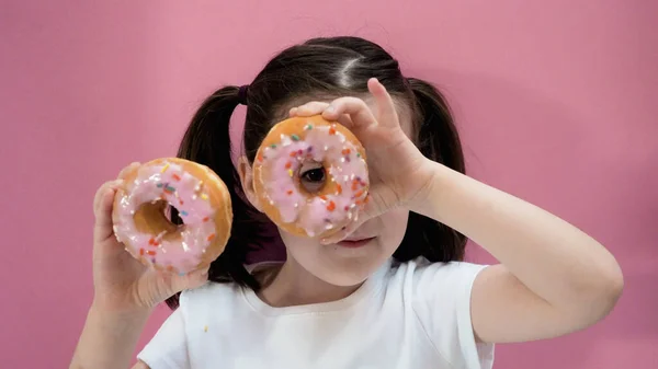 Portrait of little dark hair girl peeping through donut with icing in her hands, look at the camera. Pink background.