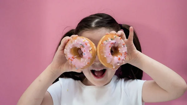 Portrait young girl makes glasses from sweet donuts with icing in her hands, smiling, looks at camera. Pink background