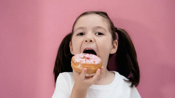 Caucasian healthy girl with ponytails eats donut opening mouth wide, healthy organic homemade food. Pink background.