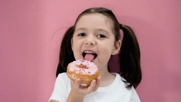 Healthy preschool girl with ponytails tongue lick pink donut and smiles. Happy toddler eats organic homemade food.