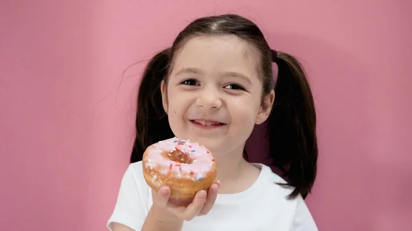 Caucasian toddler girl with ponytails holds pink donut, smiles and looks at the camera. Healthy organic homemade food