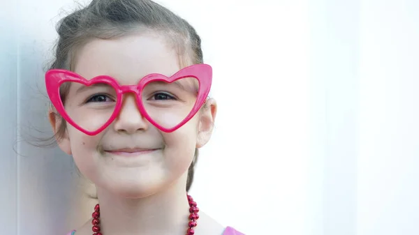 Portrait happy girl in funny red heart glasses smiles at the camera. Celebrating Valentines day. White background
