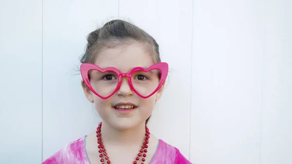 Caucasian happy toddler with funny red heart glasses looks at the camera. Celebrating Valentines day. White background