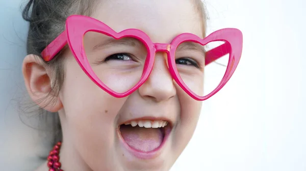 Close up. Happy brown eyes kid in red heart glasses looks at camera and smile, white background. Love and Valentines Day