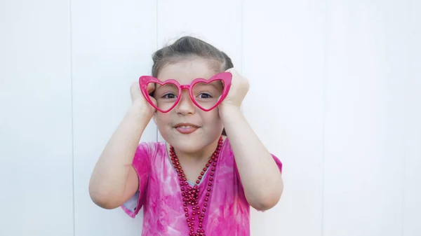 Girl in pink dress and funny red heart glasses makes face looking at camera. Celebrating Valentines day white background