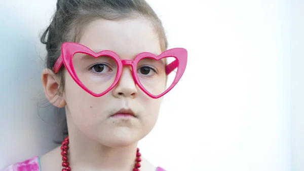Serious toddler brown eyes girl in red heart glasses looks at camera white background. Symbol of love and Valentines Day