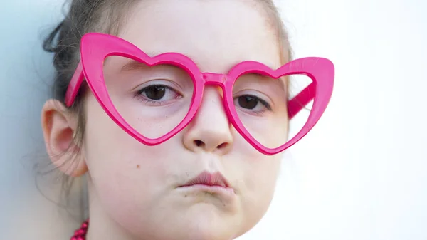 Close up. Sad brown eyes kid in red heart glasses looks at camera, white background. Love and Valentines Day