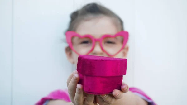 Cute toddler girl gives the red heart box present, looking at the camera in glasses symbol of love and Valentines Day